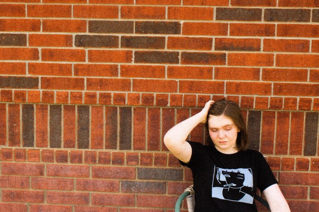 Young woman in black t-shirt sitting against a brick wall, lost in thought.